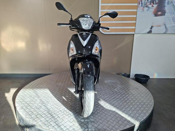 A black scooter positioned on a circular display platform, facing forward, with a minimalist background featuring neutral colors and a subtle texture.