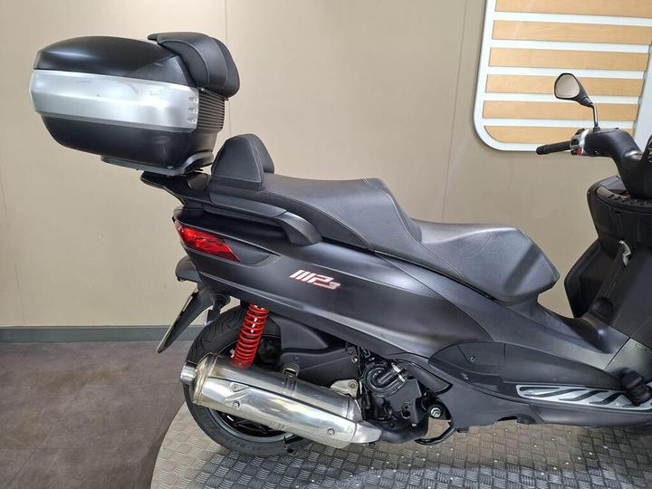 A black scooter with a silver top case, featuring a sleek design and red shock absorbers, positioned in a well-lit indoor setting with a textured floor and a light-colored wall.