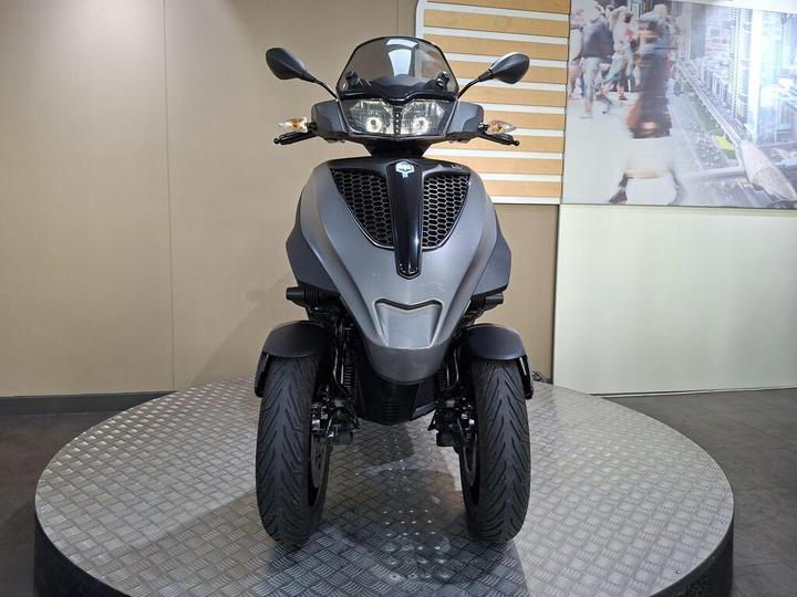 A front view of a modern gray three-wheeled scooter displayed on a circular platform, with a neutral background featuring a busy urban scene.