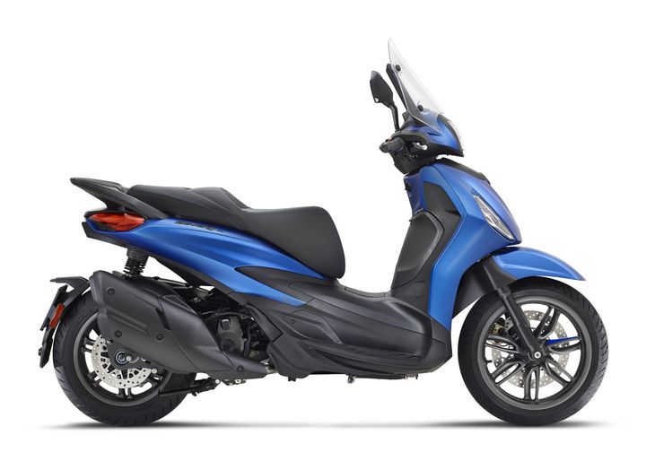 A blue scooter positioned at an angle, showcasing its aerodynamic design, seat, and front windshield. The scooter features a black undercarriage and sleek silver wheels with disc brakes.