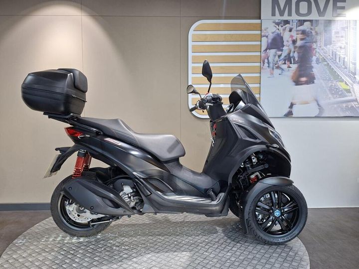Piaggio MP3 300 HPE Sport (2019) A black three-wheeled scooter with a rear storage box, displayed against a neutral background featuring a graphic with pedestrians and a modern cityscape.