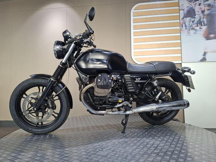 A side view of a black Moto Guzzi V7 motorcycle displayed on a textured platform, with a minimalist background featuring soft lighting.