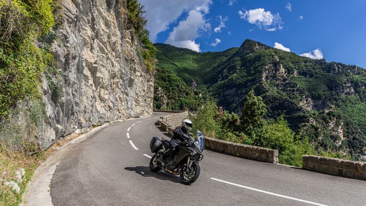 A motorcyclist in black gear rides along a winding mountain road, flanked by steep rocky cliffs and lush greenery under a partly cloudy sky.