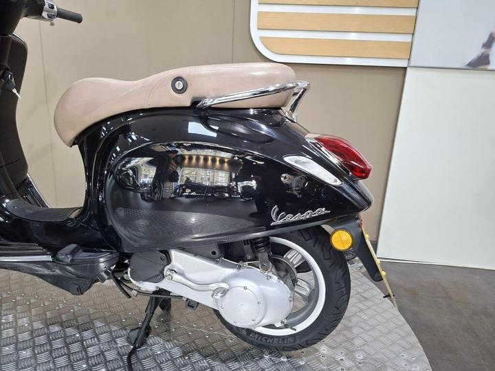 A close-up view of the rear side of a black Vespa scooter, featuring a beige seat and a glossy finish on the body, with the word "Vespa" visible on the side. The background is a neutral-toned wall and a textured floor resembling metal.