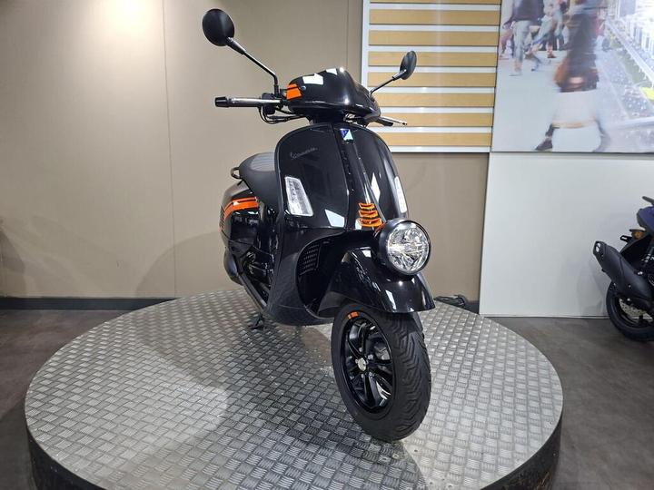 A black scooter with orange accents showcased on a circular metal platform. The scooter features a modern design with a rounded headlight and a sleek body. A light-colored wall with a display of people walking is visible in the background.