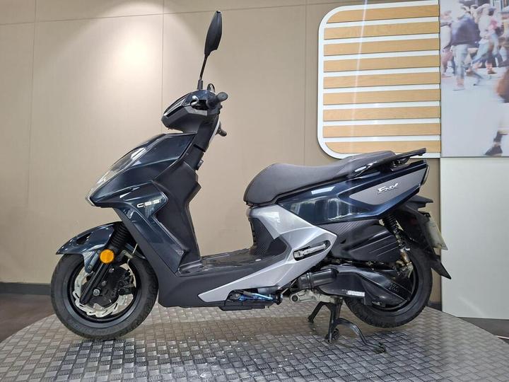 A sleek black and silver scooter is displayed on a textured platform against a neutral-colored wall, showcasing its design and features.
