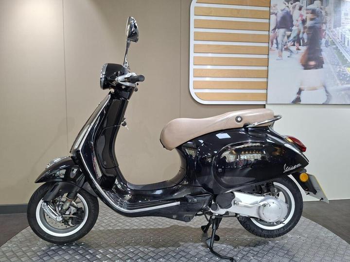 A black Vespa scooter with a beige seat is displayed against a light-colored wall, featuring a chrome rearview mirror and a patterned floor. A blurred image of people walking is visible in the background.