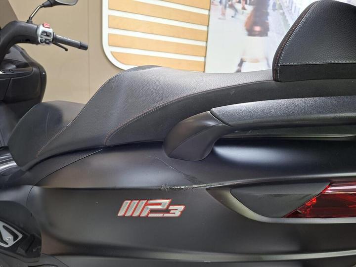 Close-up of a motorcycle seat with a textured black surface, featuring a visible scratch and the logo "MP3" in red on the side. The background includes a blurred, light-colored wall with wooden paneling.