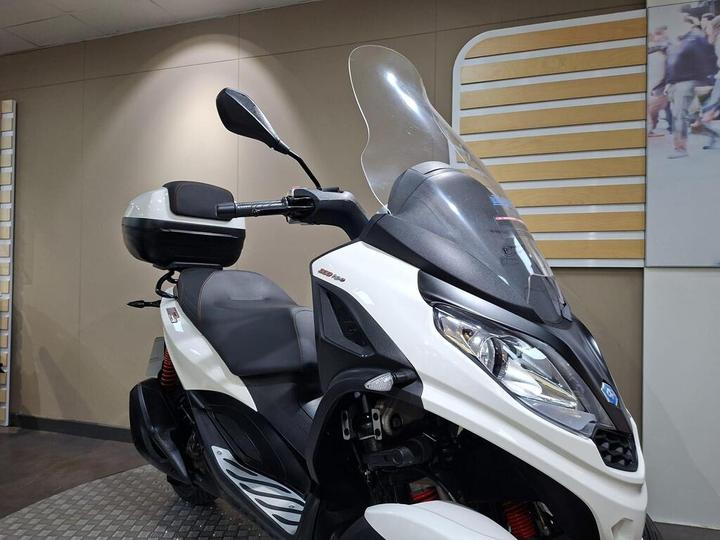 A close-up view of a white and black scooter with a large windshield, side mirror, and a storage box on the rear, set against a neutral background in a showroom.