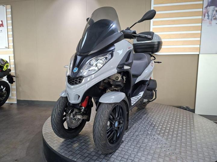 A gray three-wheeled scooter with a windshield and a storage compartment, displayed on a metal platform in a showroom.
