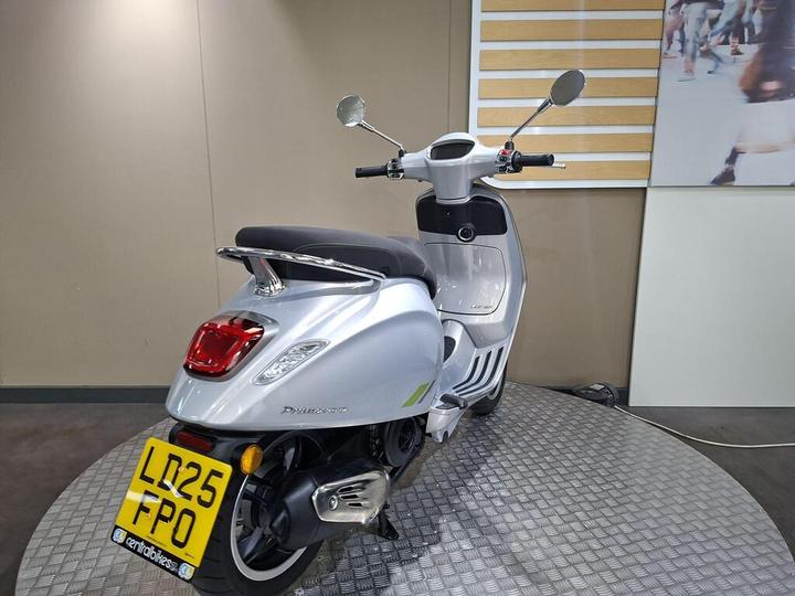 A silver scooter parked on a circular, textured platform, showcasing its rear view. The scooter's license plate is clearly visible, and a minimalist wall with simple stripes is in the background.