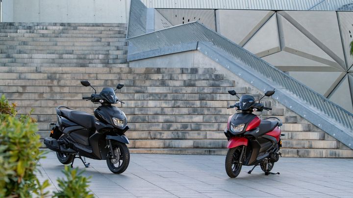 Two scooters, one black and one red, are positioned on a stone staircase outside a modern building. The black scooter is on the left, while the red scooter is on the right, both showcasing their sleek designs against the minimalist architectural backdrop.