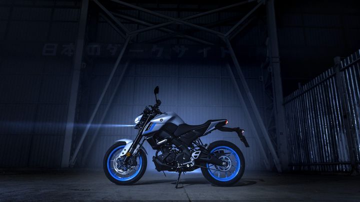 A modern motorcycle with a sleek silver body and blue accents, spotlighted in a dimly lit industrial space with metal structures in the background.