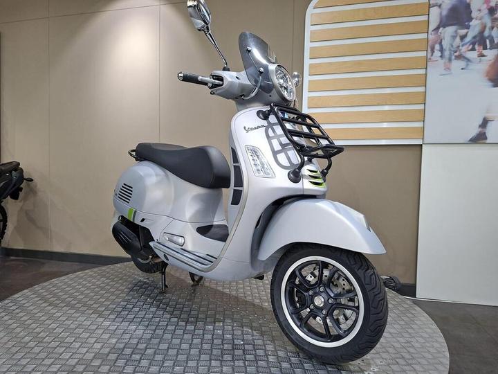 A silver scooter with a prominent front grille and distinctive wheel design, displayed in a modern showroom setting. The background features softly lit walls and promotional images, enhancing the scooter's sleek appearance.