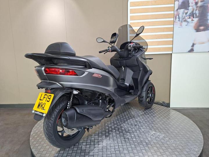 A sleek, dark gray three-wheeled scooter displayed on a circular platform, with seating for two and modern design elements, set against a neutral wall backdrop and a decorative panel.