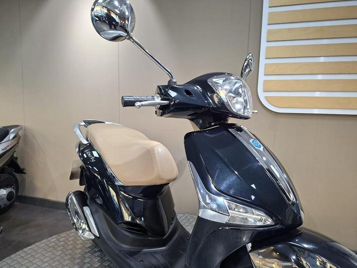 A close-up of a black scooter with a beige seat, featuring chrome mirrors and a sleek design, positioned on a textured surface against a simple, light-colored background.