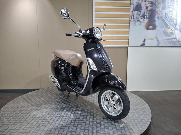 A black scooter with a brown seat displayed on a circular metal platform, with a minimalistic backdrop.