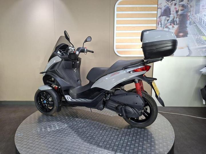 A grey three-wheeled scooter displayed on a textured surface, featuring a rear storage compartment, sporty design, and modern dashboard, set against a minimalist backdrop with a blurred urban scene.