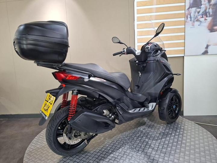 A black three-wheeled scooter with a large rear storage box, featuring red suspension springs and a sleek design, displayed on a textured circular platform against a light-colored wall.