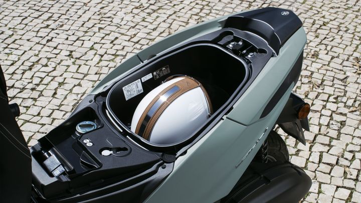An open scooter storage compartment revealing a white helmet with brown accents, against a background of cobblestone pavement.