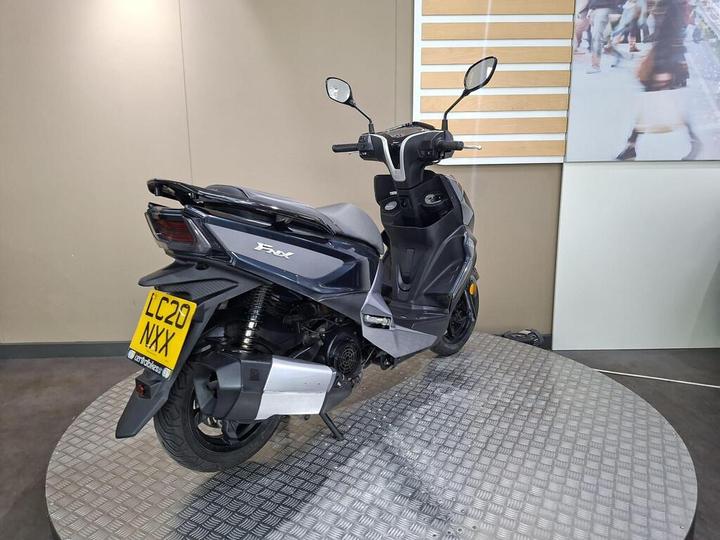 A dark gray scooter, viewed from the rear, displayed on a circular platform with a textured surface. The scooter features a license plate reading "LC20 NXX" and is set against a simple, neutral background with a partially visible image of pedestrians.