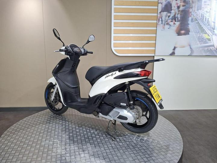 A white and black scooter displayed on a circular metallic surface, featuring a sleek design and chrome accents, with a blurred background showcasing people in an urban setting.
