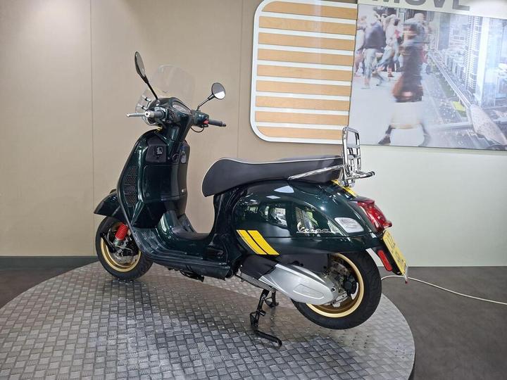 A green vintage-style scooter with a windshield and chrome accents is displayed on a circular metallic platform. The background features a wall with the word "MOVE" and an urban scene image.