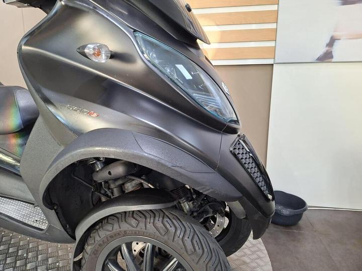A close-up view of a black three-wheeled scooter with a sleek design, showcasing its front, wheel, and distinctive headlight. The background features a neutral-colored wall with wooden panels.