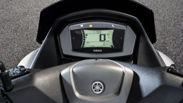 A close-up view of the dashboard of a Yamaha scooter, displaying an LCD screen with various indicators, including speed, fuel level, and odometer readings, against a textured gray background.