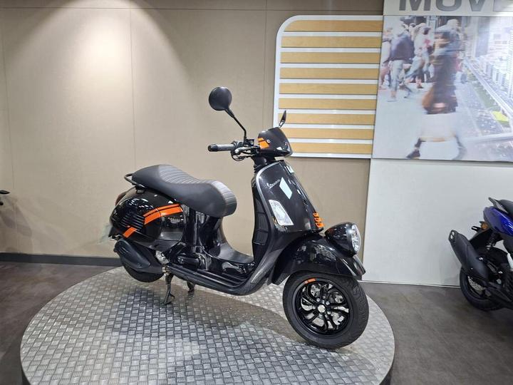 A sleek black scooter with orange accents is displayed on a round platform in a well-lit showroom, with a neutral background featuring a promotional image.