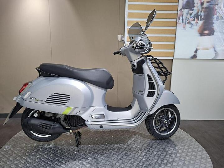 A silver scooter with a sleek design, featuring a modern vintage style, is displayed on a circular platform against a neutral background. The scooter has a black seat, visible handlebars, and a windshield.