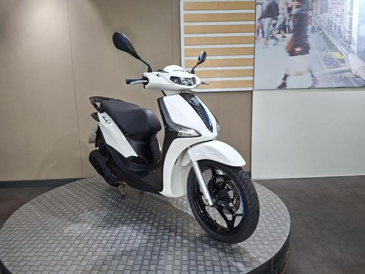 A sleek white scooter displayed on a circular metal platform, showcasing its modern design and features, with a light-colored wall and a colorful backdrop of urban scenes behind it.