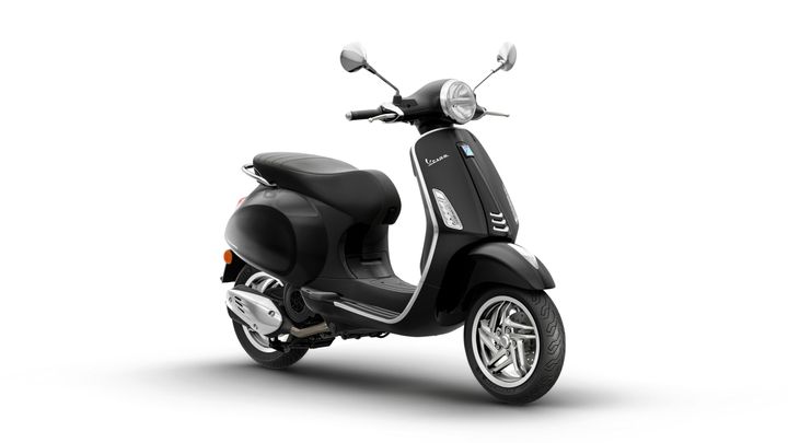 A black Vespa scooter is positioned at a slight angle, showcasing its sleek design, chrome accents, and comfortable seating. The scooter has a round headlight, mirrors, and a silver exhaust pipe, with a clean white background.