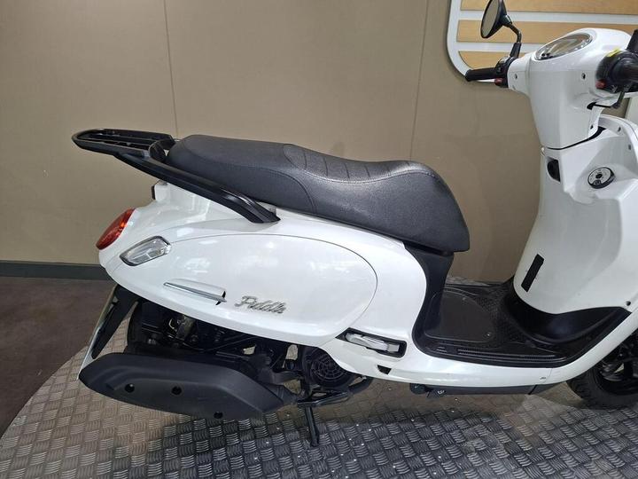 A white scooter stands on a textured surface, showcasing its sleek design, with a black seat and rear luggage rack. The scooter features a streamlined body and a modern aesthetic.