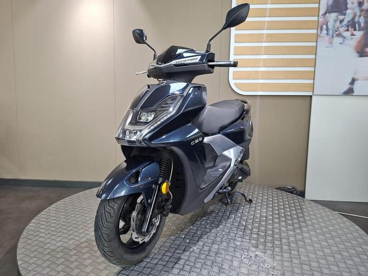 A dark blue scooter displayed on a circular platform, showcasing its sleek design and modern features, set against a neutral backdrop.