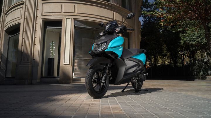 A Yamaha scooter with a black and turquoise design parked on a cobblestone street near a modern building, featuring large glass windows and trees in the background.