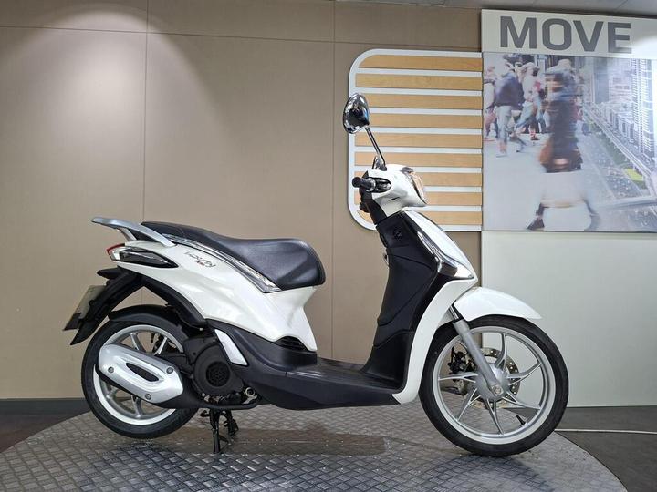 Piaggio Liberty 125 2018 A white scooter displayed at an indoor showroom against a neutral background with a modern design. The scooter features a sleek design, a comfortable seat, and a clear headlight.