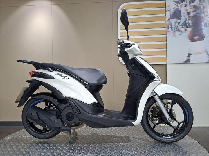 Piaggio Liberty S 125 (2025) A side view of a white scooter with black accents, featuring a sleek design, visible wheels, and a modern seat, displayed on a textured surface against a neutral background.