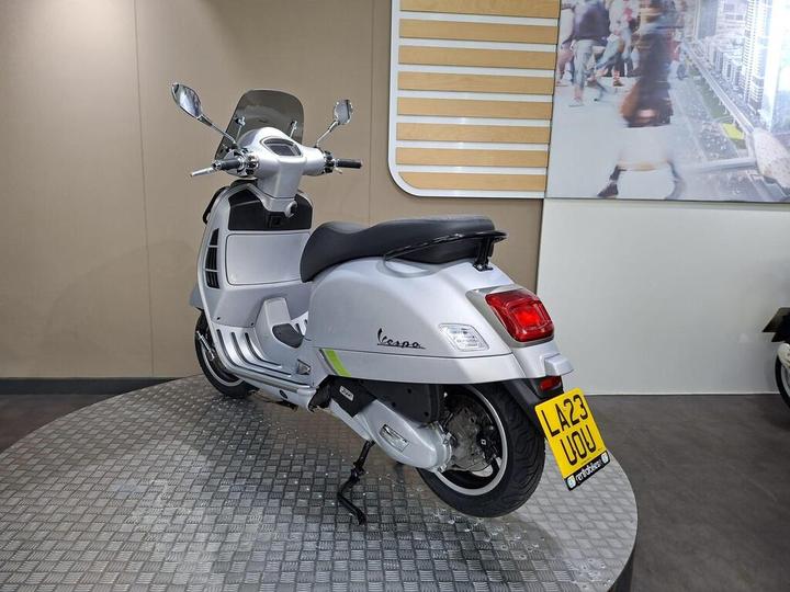 A silver scooter displayed on a circular platform with a modern backdrop, featuring a windshield, dual mirrors, and a prominent license plate reading "LA23 UOU."