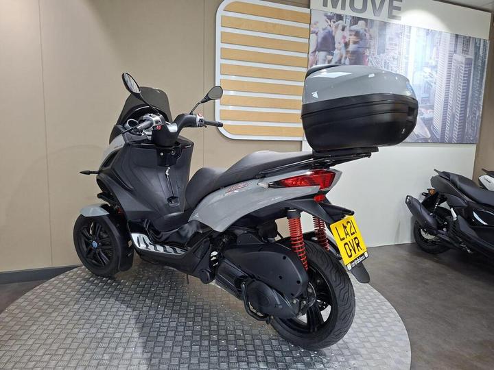 A gray scooter with a top case is displayed on a circular metallic platform against a neutral wall, featuring a "MOVE" sign and a cityscape background. The scooter shows a sleek design with prominent red suspension and black wheels.