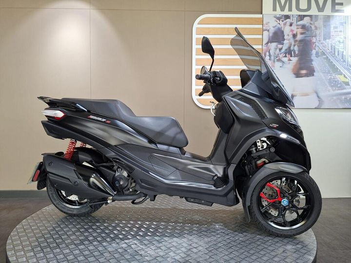 Piaggio MP3 400 (2022) A side view of a sleek, modern black three-wheeled scooter displayed on a circular metal platform, featuring red suspension and accents, set against a minimalistic interior backdrop.
