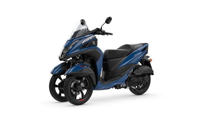 A blue Yamaha Tricity three-wheeled scooter is shown at a slight angle, featuring a sleek design, a windshield, and comfortable seating. The front wheels are larger, with a sporty look, while the body has smooth contours and a modern style.