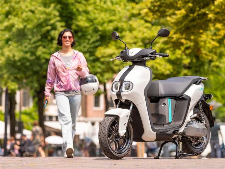 A smiling woman wearing sunglasses and a pink hoodie walks towards a white electric scooter parked on a sunny street lined with trees. She is holding a helmet and a phone.