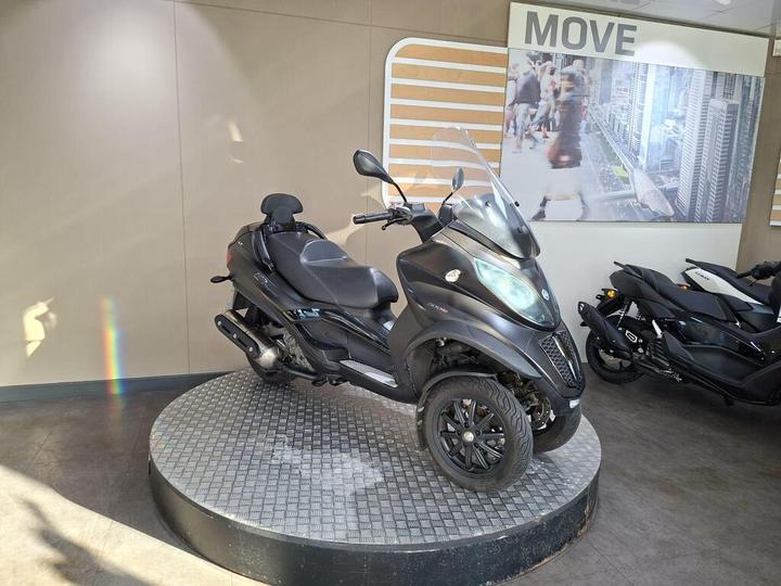 A black three-wheeled scooter displayed on a circular platform in a modern showroom, with a backdrop featuring a cityscape and the word "MOVE."