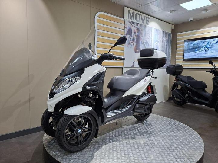 A three-wheeled scooter displayed on a circular platform in a showroom, featuring a white and black design, windshield, and a rear storage compartment. The background includes promotional graphics and a television screen showing a motorcycle image.