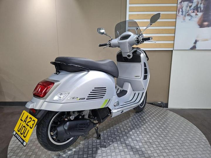 A silver scooter, featuring a streamlined design with a windscreen, is positioned on a circular metal display platform against a neutral background. The scooter's rear showcases its distinctive tail light and engine components.