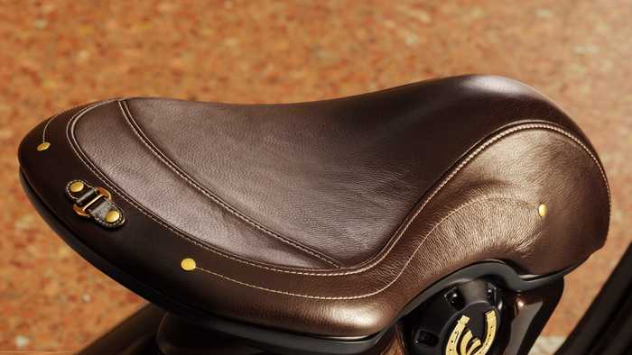 A close-up of a brown leather scooter seat with gold accents and stitching, mounted on a glossy brown scooter body. The background features a textured, warm-toned surface.