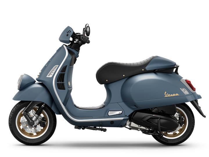 A blue Vespa scooter with a sleek design, featuring a black seat, gold-accented wheels, and a modern headlight. The scooter is positioned in profile against a white background.