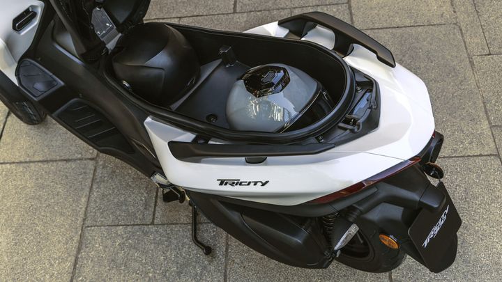 A close-up view of a white three-wheeled scooter with a helmet stored under the seat, showcasing the "Tricity" logo. The background features a textured pavement.