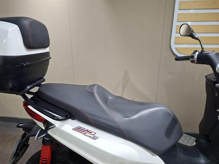 A close-up view of a scooter's seat and rear storage box, showing a sleek design with contrasting black and red stitching on the seat and a glossy white exterior.